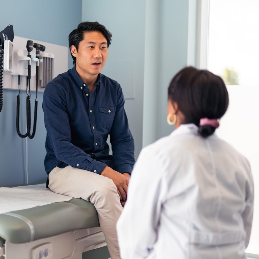 Young Asian man wearing a navy blue shirt and khaki pants having a conversation with his female physician during a primary care appointment